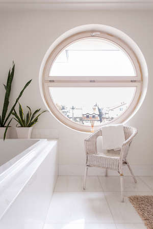 Spacious bathroom with white tiling, bathtub, chair and round windowの写真素材