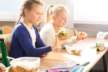 Two little pretty girls eating together healthy sandwiches during lunch break at schoolの写真素材