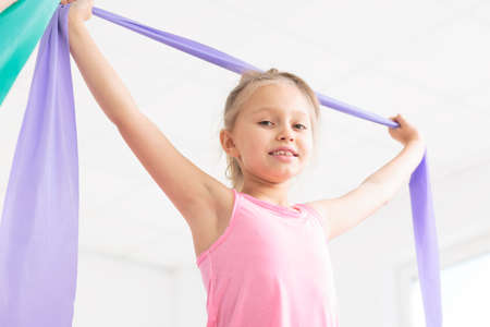 Close-up of a smiling little girl exercising with a violet resistance band in a very bright room