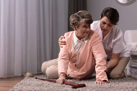 Carer helping senior woman with walking stick to stand up from floor.
