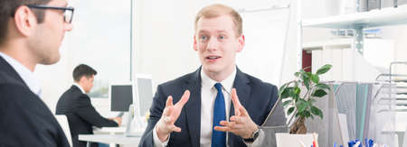 Panoramic photo of a young, smartly-dressed man in an office, having a lively discussion with his colleagueの写真素材