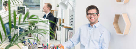 Panoramic photo of a young, smiling designer in a modern, bright office, with his colleagues talking in the backgroundの写真素材