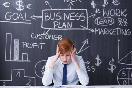 Exhausted ginger woman sitting beside desk, holding her head, business plan drawn on a blackboard in the backgroundの写真素材