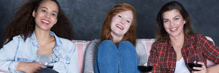 Three friends sitting on a sofa at evening, smiling and drinking wine, panoramaの写真素材