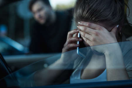 Shot of a young woman talking on the phone and covering her face and a man standing next to the carの写真素材
