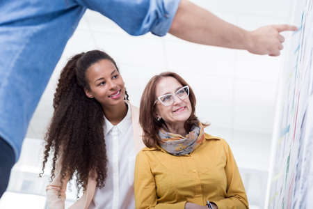 Close-up of two women in a bright office looking at some graphs to which a man is pointing in the blurred foregroundの写真素材