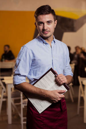 Young waiter in restaurant interior taking the menu in his hands, with restaurant tables and chairs in the backgroundの写真素材