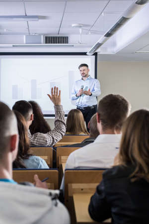 Lecture room filled with students, with an academic teacher asking questions next to bar graphs...