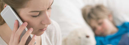 Worried woman talking on a cellphone, in the background blurred image of a small boy lying in bedの写真素材