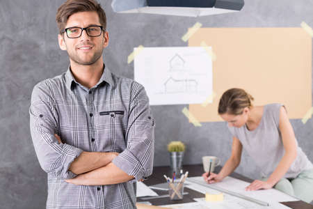 Handsome  young architect, in the background woman sitting beside desk drawing a projectの写真素材