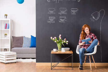 Woman sitting on a chair beside small table, reading a book, in the background blackboard coffee menu and light room with sofa and regaleの写真素材