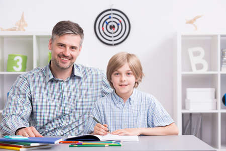Happy father and son sitting beside desk in light room, doing homeworkの写真素材