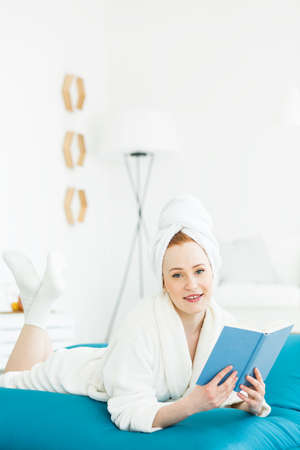 Portrait of a red-haired woman in nightgown, with a towel on her head, reading a book on a large blue sit sackの写真素材