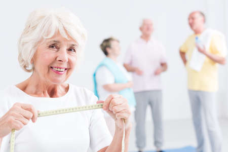 Happy senior woman smiling, holding tape measure, elderly people in the backgroundの写真素材