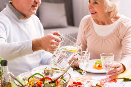 Aged happy couple eating dinner together. Man pouring cold water to the glass