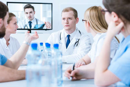 Young doctors sitting around a conference table and communicating on-line with a doctor displayed on a tv screenの写真素材