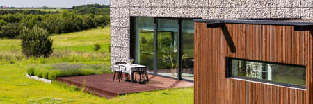 Housefront of a stone house with wooden elements and open terrace with table and chairs surrounded...