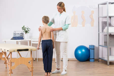 Doctor correcting small boy's body posture, standing in well equipped physiotherapist officeの写真素材