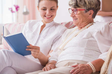 Smiled nurse and senior woman sitting together on a couch and reading a bookの写真素材