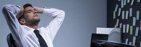 Tired businessman holding his head, sitting at office at night, panoramaの写真素材