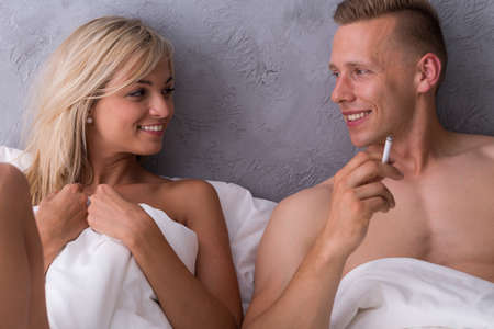 Happy blonde beauty and handsome man with a cigarette lying together in a hotel bedの写真素材