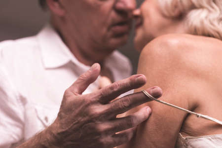 Elder couple bodies close to each other with the man's hand stripping shoulder-strap of woman's...