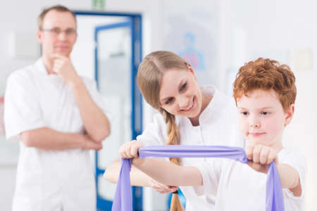 Ginger boy exercising with resistance band and two professional physiotherapistの写真素材