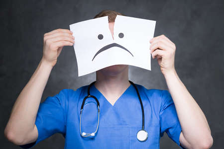 Young doctor with the stethoscope on his neck, holdling the piece of paper with sad mien covering his faceの写真素材