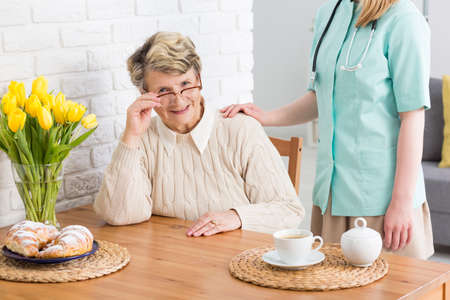 Senior woman with glasses sitting at a wooden table with cup of coffee, croissants and vase of yellow tulips, close to the nurse standing close to herの写真素材