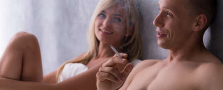 Man is smoking a cigarette and smiled woman is looking at him while they are resting in bedの写真素材