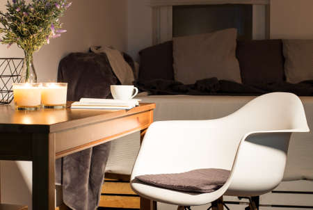 Bedroom interior with wooden table with cup of coffee and candles on, minimalistic white chair and...