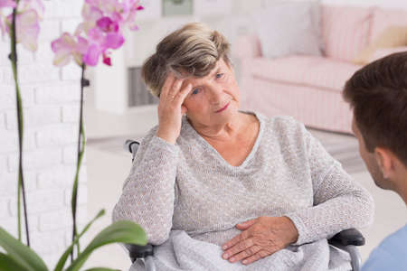 Senior woman on a wheelchair with a headache and her carer, light interiorの写真素材