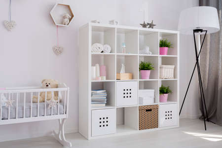 Bright baby girl room with white book case, baby cradle with a teddy bear and a lampの写真素材
