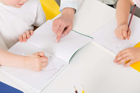 Close-up of a school desk with pupils writing simple equations in copybooks, assisted by a teacher