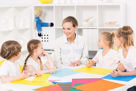 Pre-school children sitting around a table filled with coloured cutting paper, talking to their teacherの写真素材
