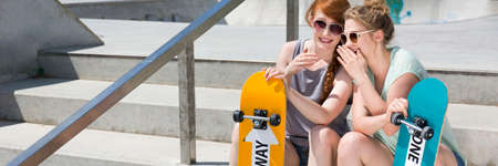 Two skater girls sitting on a city stairs with their skateboards during summer day, panoramaの写真素材