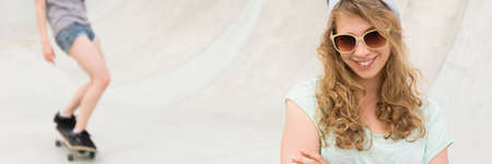 Happy girl wearing sunglasses and beanie hat, skater in the background, panoramaの写真素材