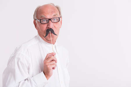 Portrait of a elderly man wearing glasses, holding a paper mustache on a stickの写真素材