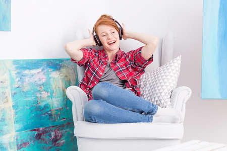 Close-up of a young woman with closed eyes, listening to music in a white armchair among blue paintingsの写真素材