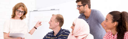 Panoramic shot of a smiling teacher pointing at a board, multicultural language class watching at...