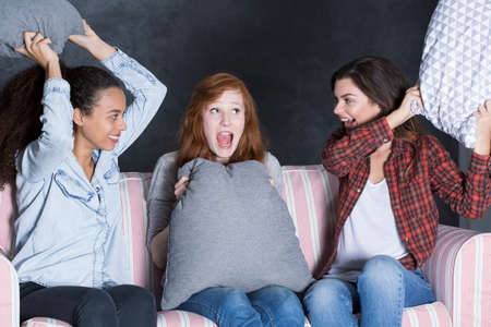 Shot of three happy friends sitting on a sofa and having a pillow fightの写真素材