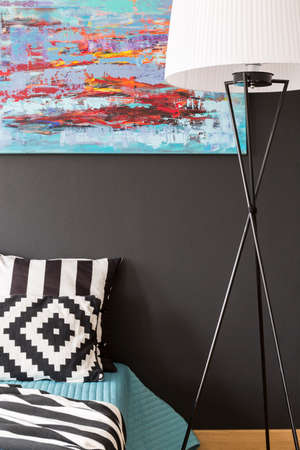 Close up of a bedroom with pattern bedding, floor lamp and modern picture on a black wall