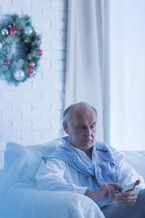 Lonely senior man sitting on sofa with cellphone during christmasの写真素材