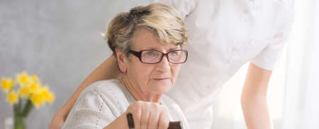 Elderly woman with glasses and hand on a cane, sitting in her home with the caregiver standing behind herの写真素材