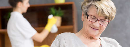 Elderly woman with glasses sitting in her home with the nurse helping her to keep the room cleanの写真素材