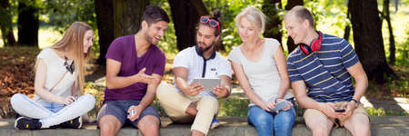 Young relaxed friends are sitting with tablet in parkの写真素材