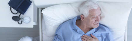 Elderly man suffering from chest pain lying in hospital bed with pressure gauge on a nightstandの写真素材