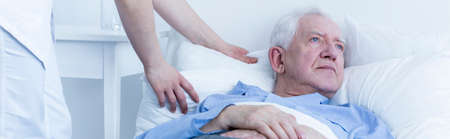 Older sad lonely man lying in hospital bed with nurse helping himの写真素材