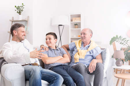 Grandfather, son and grandson sitting on sofa and talkingの写真素材