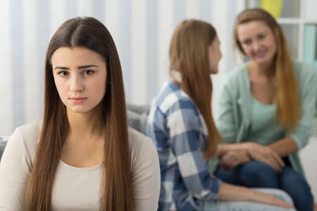 Unhappy teenage girl sitting on the couch with her friends gossiping at the backの写真素材